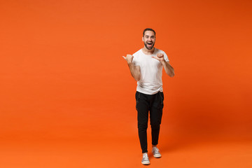 Fototapeta na wymiar Excited young man in casual white t-shirt posing isolated on bright orange wall background studio portrait. People sincere emotions lifestyle concept. Mock up copy space. Pointing thumbs aside.