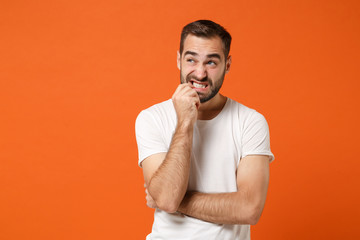 Nervous young man in casual white t-shirt posing isolated on bright orange wall background studio portrait. People sincere emotions lifestyle concept. Mock up copy space. Gnawing nails, looking up.