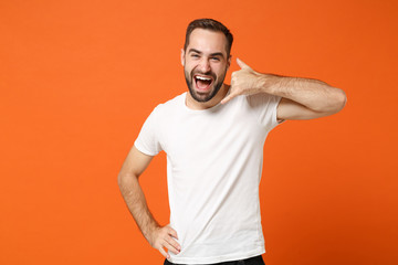 Cheerful young man in casual white t-shirt posing isolated on orange background in studio. People sincere emotions lifestyle concept. Mock up copy space. Doing phone gesture like says call me back.