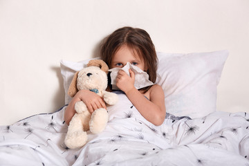 Obraz na płótnie Canvas Picture of ill little girl looking directly at camera, holding her handkerchief near nose, feeling unwell, having running nose, hugging her teddy bear, lying in her bedroom. Health problems concept.