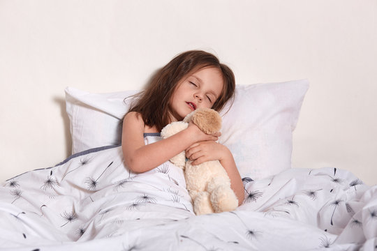 Image of charming magnetic little infant sleeping in her light bedroom alone, lying in soft cosy bed, hugging her favourite bear toy, closing eyes and opening mouth. Happy childhood concept.