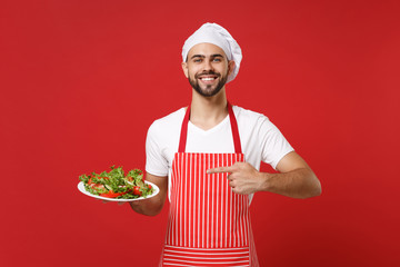 Smiling male chef cook or baker man in striped apron toque chefs hat posing isolated on red background. Cooking food concept. Mock up copy space. Pointing index finger on plate with vegetable salad.