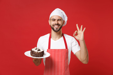 Bearded male chef cook or baker man in striped apron white t-shirt toque chefs hat posing isolated on red background. Cooking food concept. Mock up copy space. Hold plate with cake showing OK gesture.