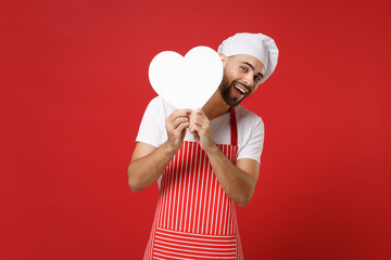 Funny young bearded male chef cook or baker man in striped apron t-shirt toque chefs hat isolated on red background. Cooking food concept. Mock up copy space. Holding in hands white empty blank heart.