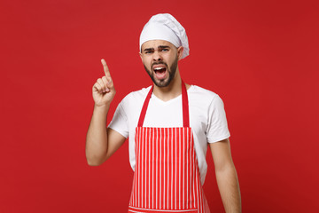 Angry bearded male chef cook or baker man in striped apron toque chefs hat posing isolated on red background. Cooking food concept. Mock up copy space. Screaming, swearing, pointing index finger up.