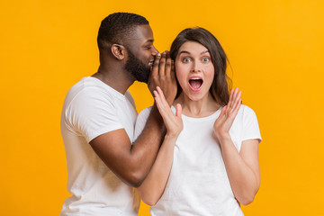 Smiling black guy whispering something into his shocked girlfriend's ear