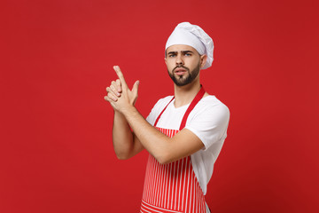 Concerned young bearded male chef cook or baker man in striped apron white t-shirt toque chefs hat posing isolated on red background. Cooking food concept. Mock up copy space. Holding hands like gun.