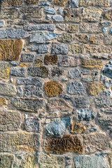 Natursteinmauer mit Steinen in mehreren Farben wie dunkelgrau, graublau, über einen dunklen, gelben Stein bis zum hellen Sandstein