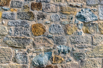 Natursteinmauer mit Steinen in mehreren Farben wie dunkelgrau, graublau, über einen dunklen, gelben Stein bis zum hellen Sandstein