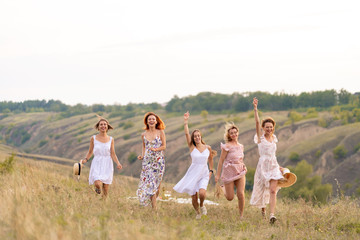 Obraz premium The company of cheerful female friends have a great time together on a picnic in a picturesque place overlooking the green hills. Girls in white dresses dancing in the field