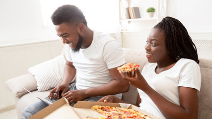Obraz na płótnie Canvas Young afro couple eating pizza and browsing movies on laptop,