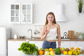 Young girl shows ok symbol in a white kitchen. Table with herbs, vegetables and fruits. Proper healthy nutrition.