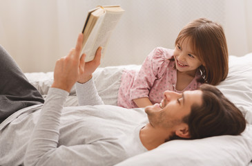 Young man reading book to his little smiling daughter