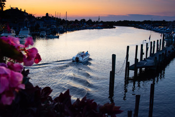 Vibrant and beautiful sunset scene of the harbor in Lewes, Delaware taken from drawbridge and featuring flowers, a single receding boat with wake, and a pier. © DanaDaglePhotography