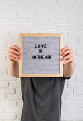 Man holding a black felt letter board with the words Love Is In The Air against the white brick wall