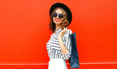 Beautiful young smiling woman with shopping bags wearing white striped shirt, black round hat on city street over red wall background