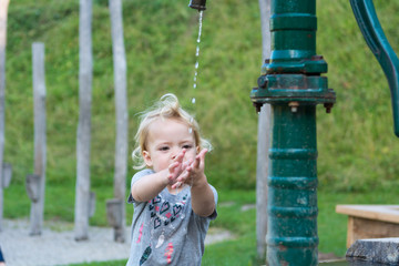 Cute blonde girl washing hands using hand water pump in local park.