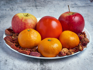 Heap of fresh seasonal apples, persimmon, mandarins and dried fruits on white plate over grey marble background. Healthy, clean eating, weight loss, dieting food concept