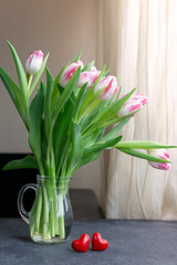 Bouquet of white with pink tulips in glass vase with two red hearts on table near window. Birthday, Love, Valentine's Day concept. Natural light, copy space. Greeting card.