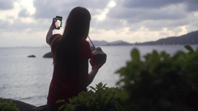 Young women silhouette taking selfies on sunset. Tropical beach view. Evening blurred ocean view. Girl drinking coconut and making photos 