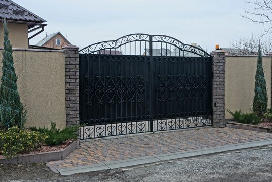 a large black iron gate with a forged pattern and part of a gray concrete fence with green decorative vegetation on the street