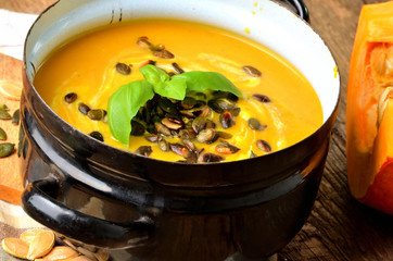 Enamel pot of cream pumpkin, carrot soup with fresh basil, pumpkin seed on rustic wooden table.