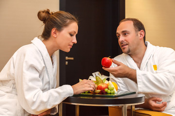 Young happy just married Caucasian couple in white bathrobes having fruits after spa on vacation. Man offers an apple to woman as Adam once tempted Eva in paradise. Temptation concept.