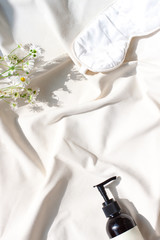 Organic cotton white bed sheets concept frame with chamomile, a sleeping mask and a body lotion cosmetic bottle . Copy space