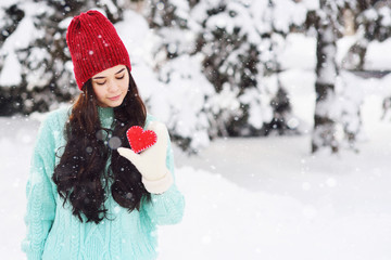 Young pretty girl in a warm blue sweater and mittens on the background of snow-covered trees holding a red heart and smiling. Valentine's day.