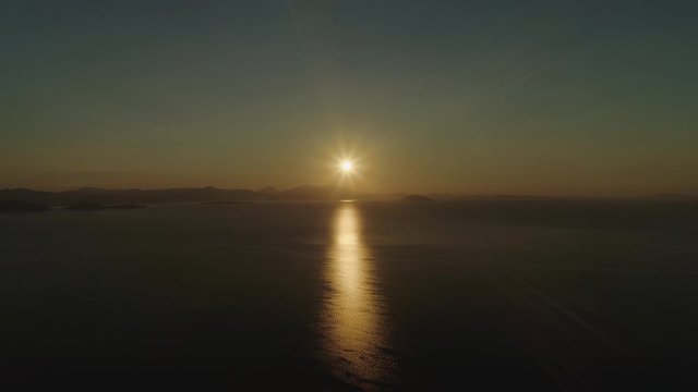 Flying high above the sea or ocean at sunset, with sun reflecting on the surface of the water.Scenic video. Mountains are visible on the horizon. Video 4 of 5.