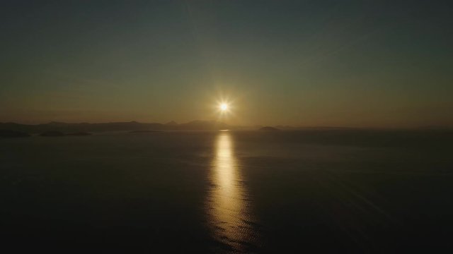 Flying high above the sea or ocean at sunset, with sun reflecting on the surface of the water.Scenic video. Mountains are visible on the horizon. Video 3 of 5.