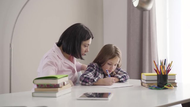 Camera approaching to tired Caucasian mother yawning as helping daughter to do homework. Cute brunette schoolgirl studying with mom at home. Support, education, intelligence.