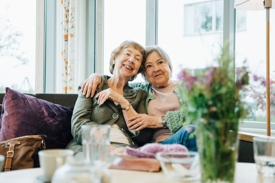 Smiling elderly women sitting with arm around on sofa against window at retirement home