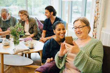 Retired elderly woman sharing smart phone with smiling young female nurse at community outreach