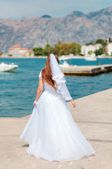 Fototapeta na wymiar Bride in white wedding dress with veil at summer sunny day, back view. Brown-haired woman walking on background of sea and mountains