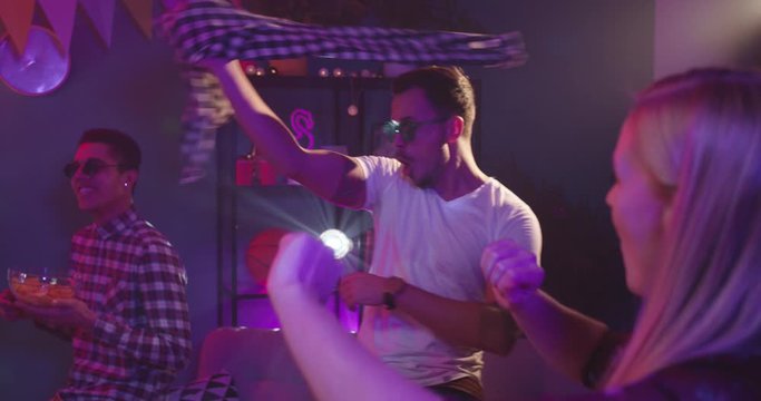Young Caucasian handsome and stylish guy in sunglasses dancing and throwing away his shirt like dancing strip at home party among friends.