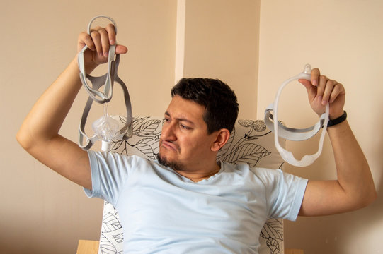 Young man is trying to choose which mask he will use for his sleep apnea disease, nasal CPAP mask or under the nose nasal CPAP mask. Man looking at nasal CPAP mask with worried face. 