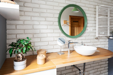 Stylish modern interior of bathroom with white brick wall and wooden counter. Ceramic wash basin ,round mirror and plant in bright bathroom in apartment.