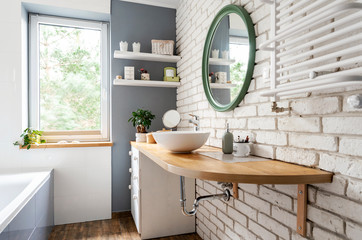 Bright interior of bathroom with window, wooden furniture and counter, round mirror and white brick wall. Simple and stylish design in scandinavian style.