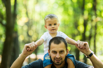 Cute little boy and his handsome young dad sitting on his father's shoulders while resting in the park.