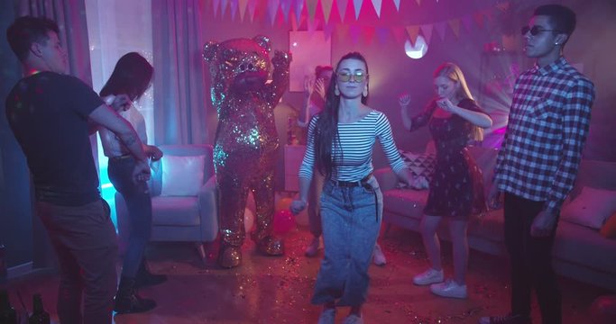 Beautiful sexy caucasian young girl in sunglasses dancing and moving with music at home party among other people and person in a costume of big golden life-size doll of teddy bear. At home party.