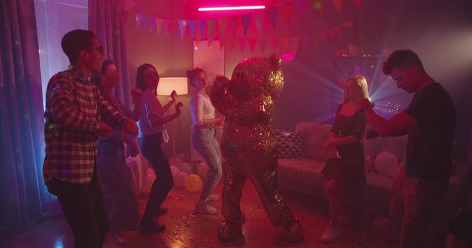 Nice home party with lights and decorations like disco - young happy guys, girls and one golden big life-size doll of teddy bear dancing together and celebrating something in the room.