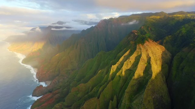Fabulous aerial view over fantastically beautiful Hawaiian coast. Setting sun dramatically illuminates bizarre mountain slopes covered with tropical vegetation. Clouds wrap around the distant peaks.4K