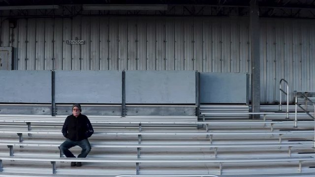 Aerial pan shot of retired coach sitting on bleachers alone
