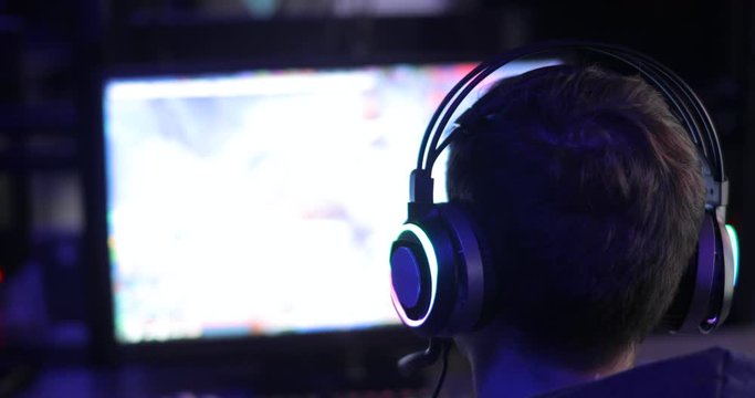 Guy in addiction. Concept of relax and dependence on computer games. Online team gaming in network. Man gamer in headset is playing video game looking at screen in dark, back view. Free time, leisure.