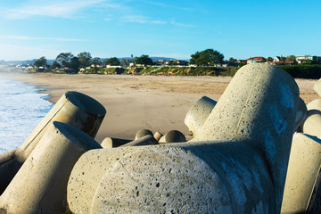 Reinforced concrete dolosse protecting breakwater jetty of Santa Cruz Harbor. Background sandy beach and blue sky - obrazy, fototapety, plakaty