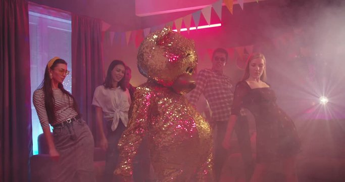 Cool funny big golden teddy bear doll in life-size dancing funny dance movement and joyful friends repeating after it on the background at home party.