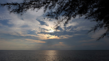 Fototapeta na wymiar Seaside with a setting sun hidden behind clouds, and silhouette of casuarina tree leaves in the foreground.