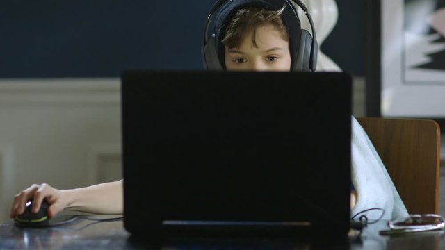 Medium shot of young boy working on a laptop with headphones on at the dining room table