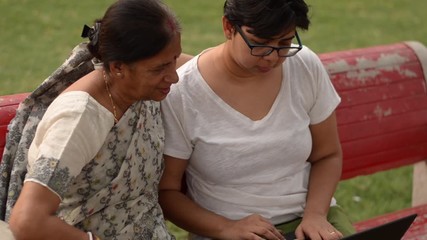 Young Indian woman helping her old Indian Bengali mother on a laptop sitting on a red bench in a park in Delhi Concept shot digital India and technology literacy for senior citizens - Powered by Adobe